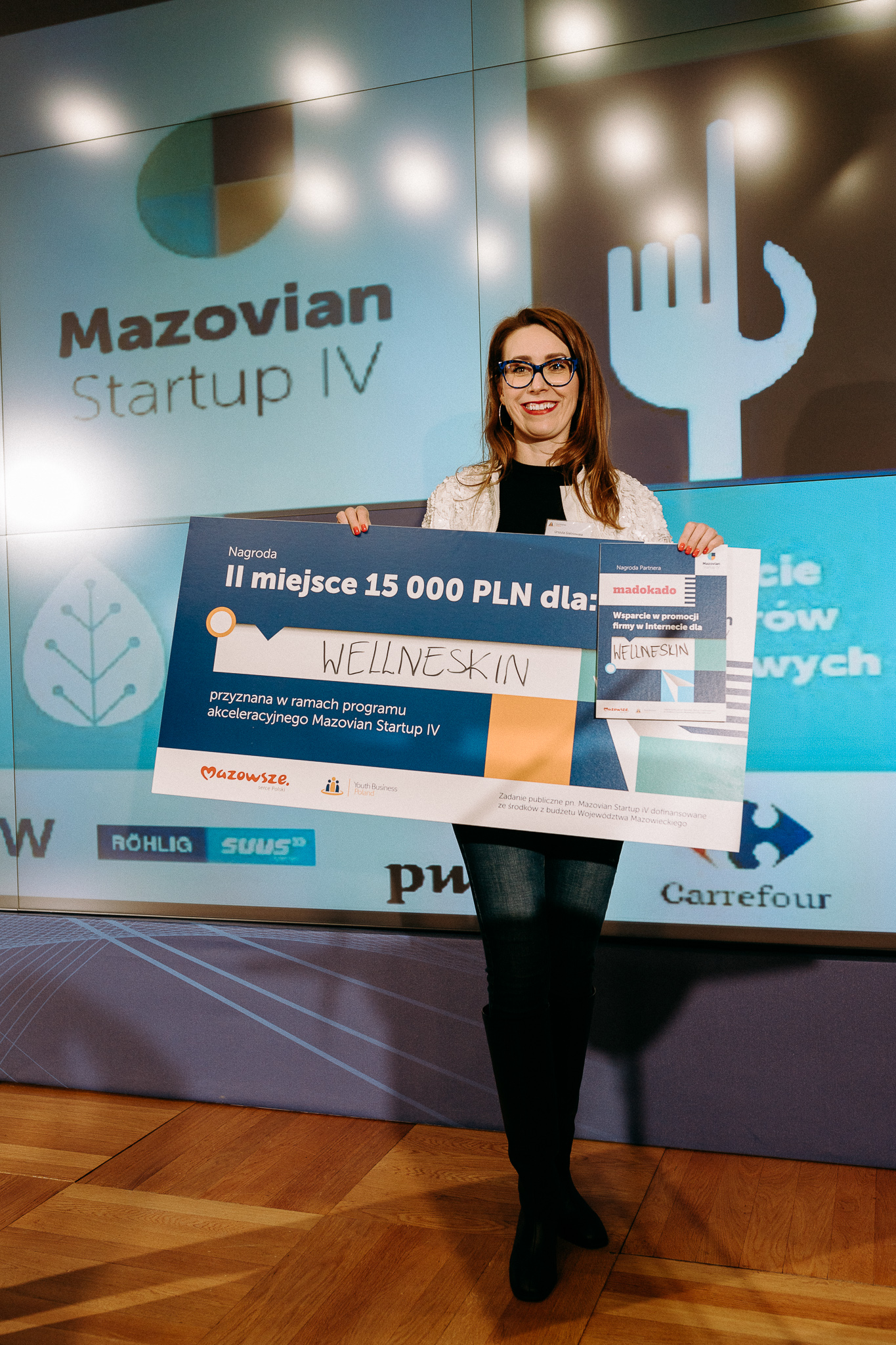Młoda usmiechnięta kobieta, trzymająca przed sobą symboliczny czek na 15 000 zł. W tle prezentacja i logo Mazovian Startup IV.