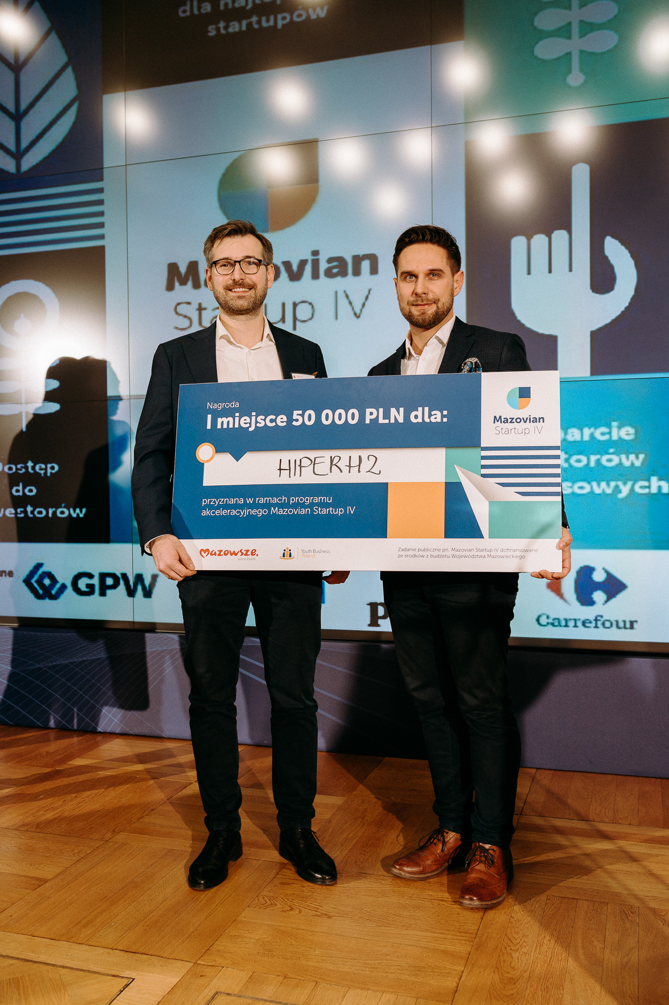 Dwóch mężczyzn, trzymają przed sobą symboliczny czek na 50 000 zł. W tle prezentacja i logo Mazovian Startup IV.